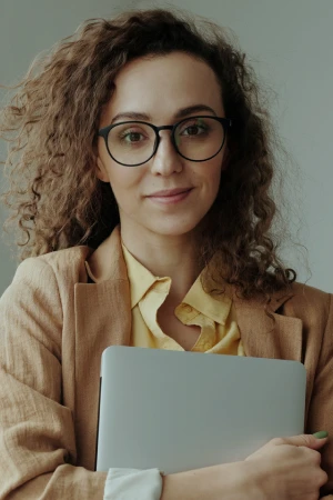 Woman with curly hair wearing glasses and a brown blazer, holding a laptop close to her chest.