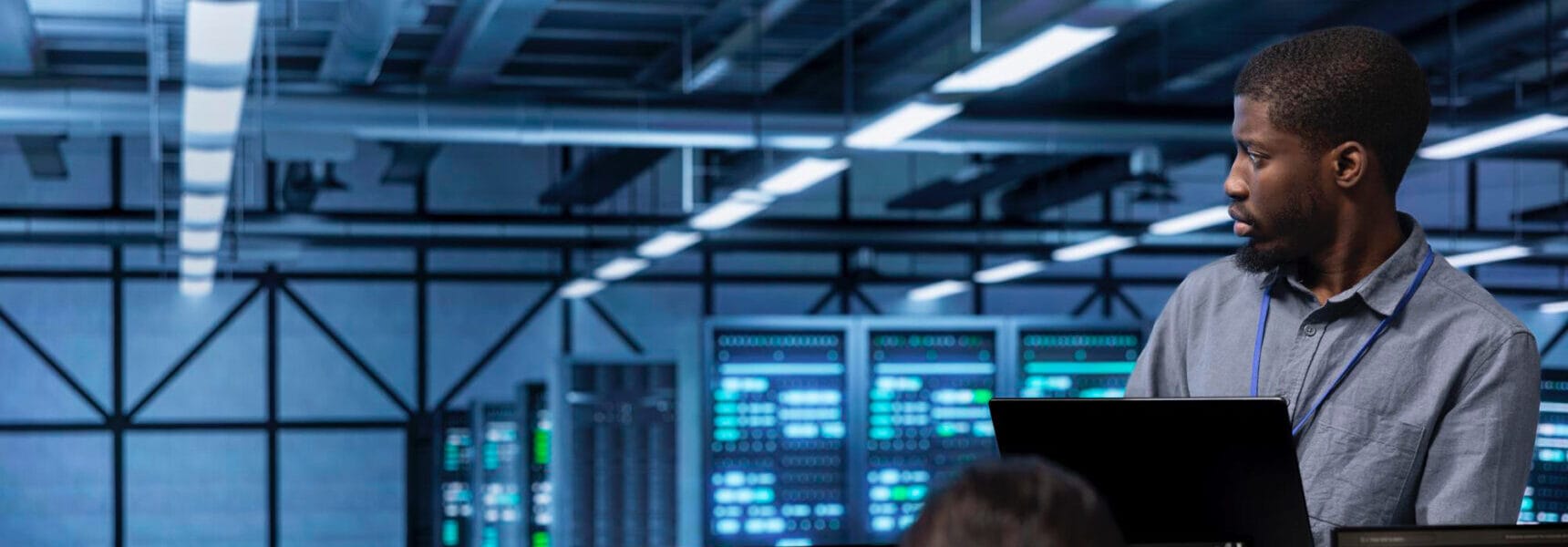 A man with short hair looking away while working on a laptop in a data center environment.