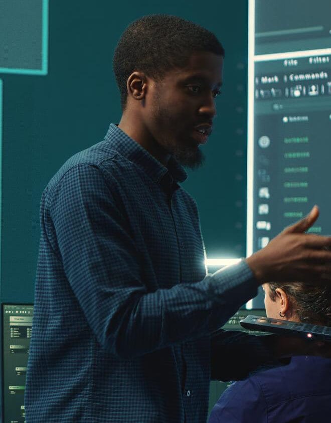 A man in a blue checkered shirt presenting in front of digital screens displaying data and code.