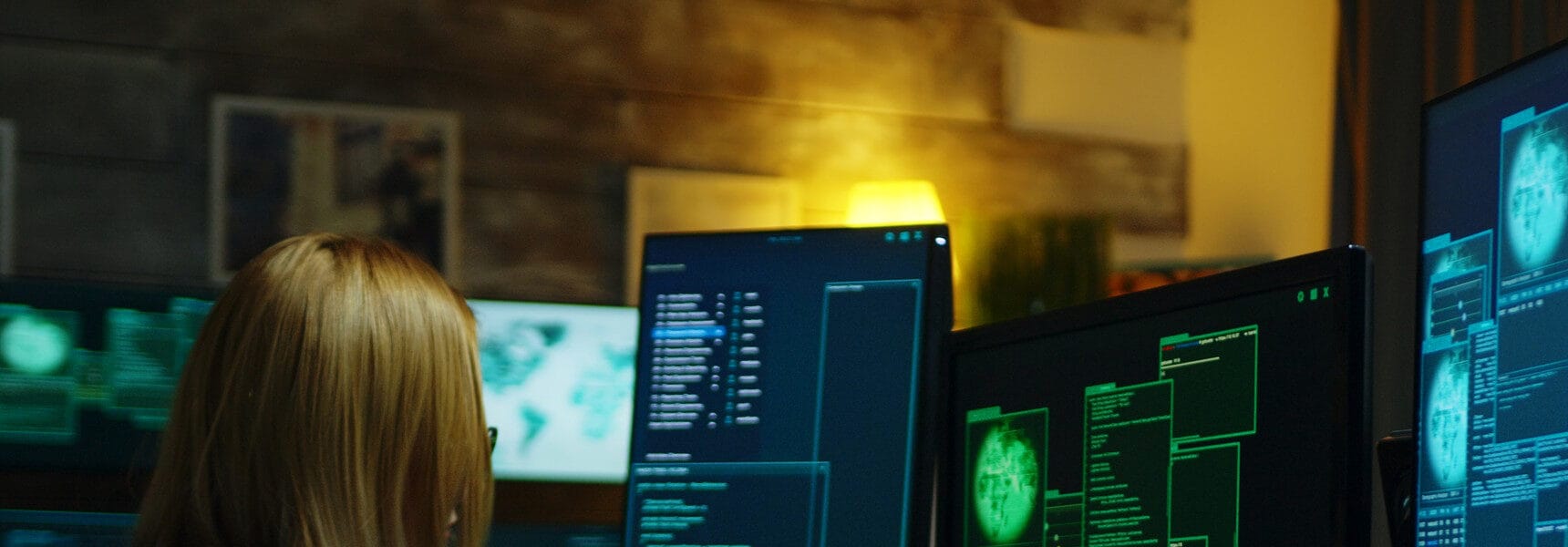 Individual with long blonde hair working at multiple computer monitors displaying various data and graphics in a dimly lit environment.