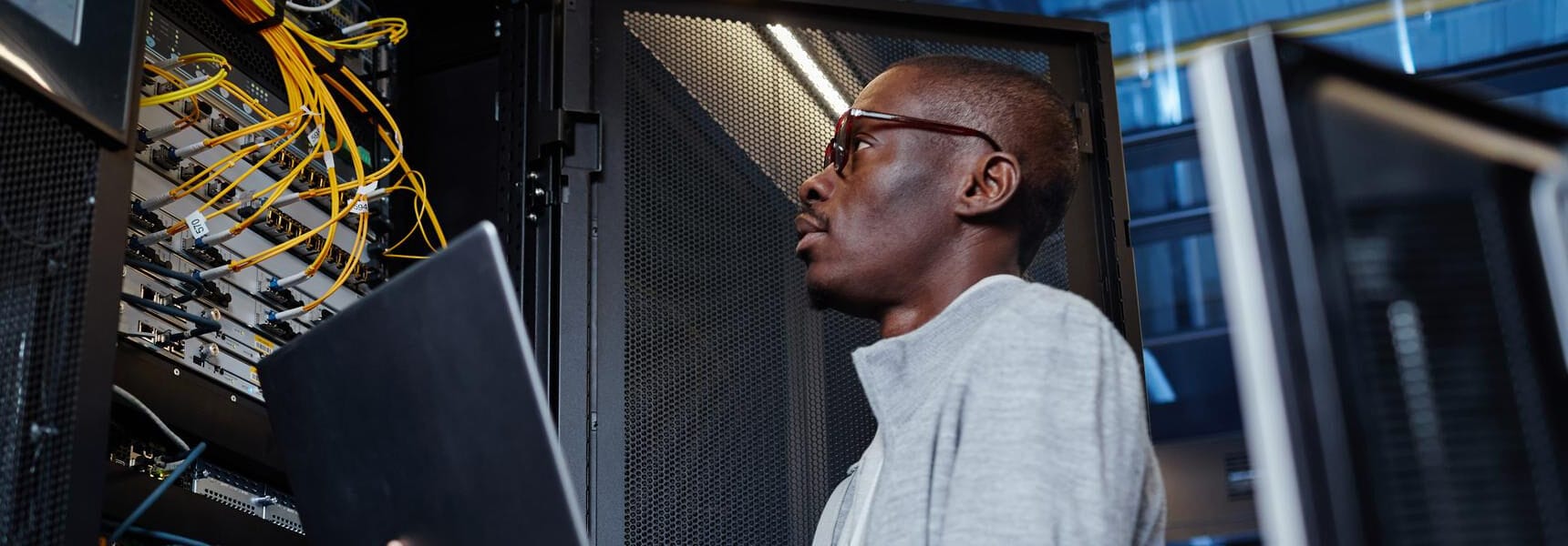 A man in glasses holding a laptop while standing near server racks with visible cables.
